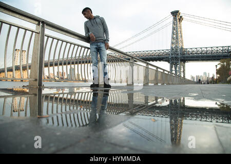 Ein junger Mann wartet entlang der East River, darunter die Williamsburg Bridge. Während des Sonnenuntergangs ein Herbst in New York City gedreht. Stockfoto