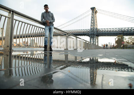 Ein junger Mann wartet entlang der East River, darunter die Williamsburg Bridge. Während des Sonnenuntergangs ein Herbst in New York City gedreht. Stockfoto