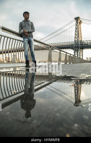 Ein junger Mann wartet entlang der East River, darunter die Williamsburg Bridge. Während des Sonnenuntergangs ein Herbst in New York City gedreht. Stockfoto