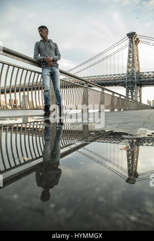 Ein junger Mann wartet entlang der East River, darunter die Williamsburg Bridge. Während des Sonnenuntergangs ein Herbst in New York City gedreht. Stockfoto