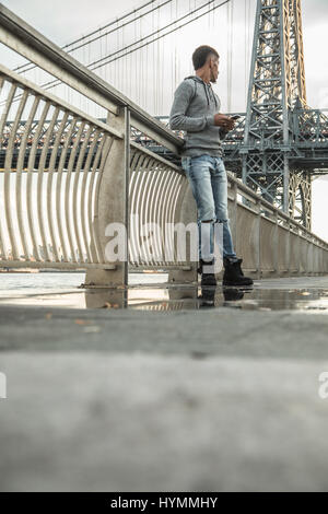 Ein junger Mann wartet entlang der East River, darunter die Williamsburg Bridge. Während des Sonnenuntergangs ein Herbst in New York City gedreht. Stockfoto