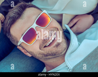 Glücklich lächelnd, bärtiger Mann mit bunten Sonnenbrille liegen auf seine Freundin. Er schaut in die Kamera Stockfoto