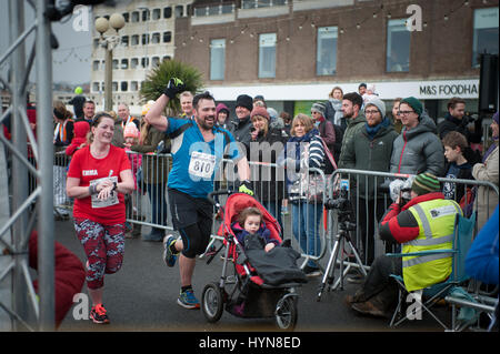 Ein Mann drückt die Kreide Buggy feiert während Überquerung der Ziellinie in Worthing Halbmarathon in Worthing, West Sussex, UK. Stockfoto