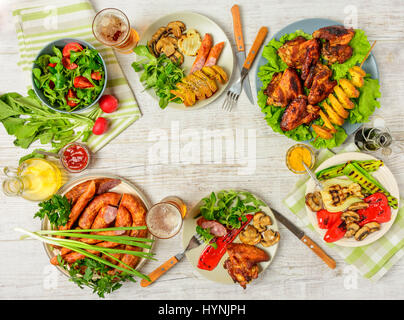 Esstisch mit verschiedene Lebensmittel, gebratene Hühnerflügel, Würstchen, gegrillte Gemüse in einen Topf geben, Salat und Lagerbier auf Holztisch. Ansicht von oben. Kopieren von spac Stockfoto