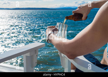 Mann sitzt am Meer Gießen Microbrew in frostigen Becher während der Sitzung in Adirondack Stühle Stockfoto