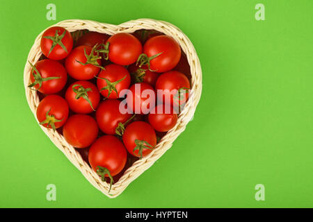 Rote, Reife frische Cherry Tomaten in kleine Herz geformten Weidenkorb auf grünem Papierhintergrund Stockfoto