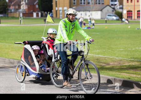 Frau Radfahrer auf Bike / Fahrrad mit + 3 Kinder; Co-Pilot Kindersitz mit Helm & Abschleppen Zyklus Chariot Anhänger mit zwei / 2 Kinder mit Helmen. VEREINIGTES KÖNIGREICH. Stockfoto