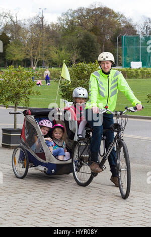 Frau Radfahrer auf Bike / Fahrrad mit + 3 Kinder; Co-Pilot Kindersitz mit Helm & Abschleppen Zyklus Chariot Anhänger mit zwei / 2 Kinder mit Helmen. VEREINIGTES KÖNIGREICH. Stockfoto