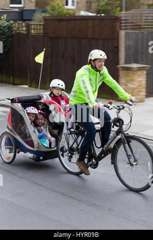 Frau Radfahrer auf Bike / Fahrrad mit + 3 Kinder; Co-Pilot Kindersitz mit Helm & Abschleppen Zyklus Chariot Anhänger mit zwei / 2 Kinder mit Helmen. VEREINIGTES KÖNIGREICH. Stockfoto