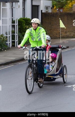 Frau Radfahrer auf Bike / Fahrrad mit + 3 Kinder; Co-Pilot Kindersitz mit Helm & Abschleppen Zyklus Chariot Anhänger mit zwei / 2 Kinder mit Helmen. VEREINIGTES KÖNIGREICH. Stockfoto