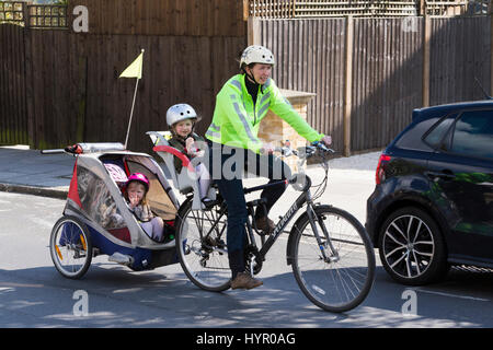 Frau Radfahrer auf Bike / Fahrrad mit + 3 Kinder; Co-Pilot Kindersitz mit Helm & Abschleppen Zyklus Chariot Anhänger mit zwei / 2 Kinder mit Helmen. VEREINIGTES KÖNIGREICH. Stockfoto