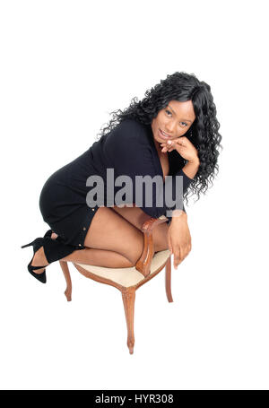 Eine schöne Afroamerikaner, kniend auf einem alten Sessel in einem schwarzen Kleid und high Heels, für weißen Hintergrund isoliert. Stockfoto