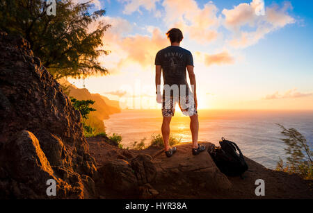 Ein männlicher Wanderer schaut auf den Sonnenuntergang an einem Kauii Wanderweg mit Blick auf den Sonnenuntergang über dem Pazifischen Ozean. Stockfoto