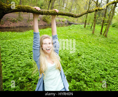 Porträt einer schönen jungen Frau lächelnd und hängen an einem Ast von einem Baum im Wald Stockfoto