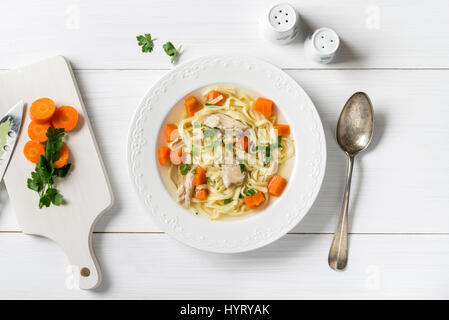 Frische Hühnersuppe mit Gemüse und Nudeln in eine Schüssel mit Möhren und Petersilie auf weißen Tisch, Ansicht von oben Stockfoto