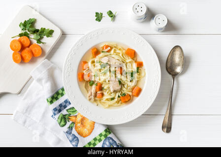 Frische Hühnersuppe mit Gemüse und Nudeln in eine Schüssel mit Möhren und Petersilie auf weißen Tisch, Ansicht von oben Stockfoto