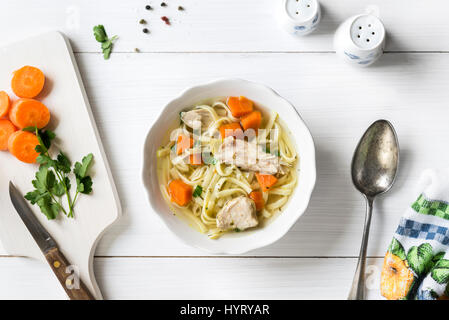 Frische Hühnersuppe mit Gemüse und Nudeln in eine Schüssel mit Möhren und Petersilie auf weißen Tisch, Ansicht von oben Stockfoto