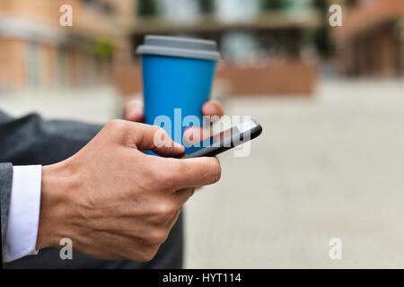 Nahaufnahme eines jungen kaukasischen Geschäftsmann in einem grauen Anzug mit einem Smartphone und einem Kaffeetrinken im freien Stockfoto