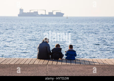 THESSALONIKI, Griechenland - 25. Dezember 2015: Oma und ihre Enkel Ruhe direkt am Meer, eine Frachtschiff sehen Sie im Hintergrund Pictu Stockfoto