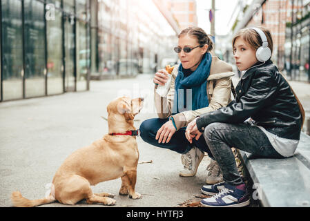 Mutter Sandwich zu essen und Blick auf einen Hund während Tochter anhören von Musik auf der Straße Stockfoto