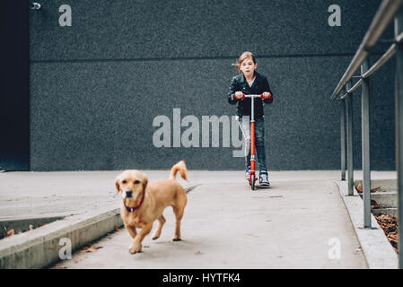 Mädchen tragen schwarze Leder Jacke treibende roten Roller mit einem kleinen gelben Hund im Vordergrund Stockfoto