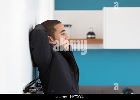 Seitenansicht der Mann im schwarzen Mantel stützte sich auf weiße Wand mit Armen hinter Kopf, Blick geradeaus und entspannen Stockfoto