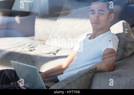 Nachdenklicher junger schwarzer Mann im weißen T-shirt auf grauen Sofa holding Laptop und unglücklich wegsehen drinnen sitzen Stockfoto