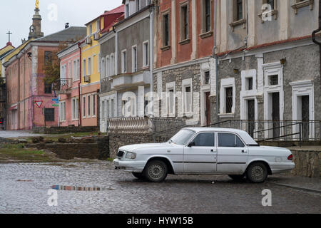 Alte weiße Volga Auto geparkt auf einer gepflasterten Seitenstraße in Kamenez-Podolsk, Westukraine. Die Straße ist noch nass vom letzten Regen Stockfoto