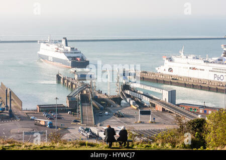 England, Dover. Vordergrund Edlerly Paar sitzt auf der Bank auf einer Klippe mit Blick auf die Fähre Terminal mit Fähren angedockt und Segeln im Hintergrund. Ansicht von hinten Paar, ferne erschossen. Stockfoto