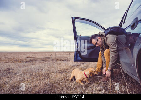 Wanderer im Auto zu sitzen und die Vorbereitung ihrer Schuhe neben ihm ist ein kleiner gelber Hund Stockfoto
