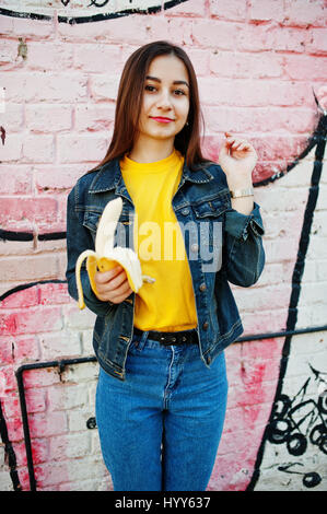 Schöne Teenager-Mädchen essen Bananen, gelbes T-shirt, Jeans in der Nähe von Graffitiwand tragen. Stockfoto