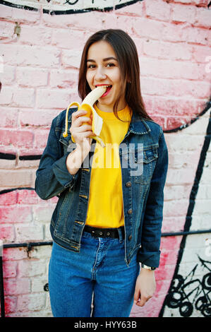 Schöne Teenager-Mädchen essen Bananen, gelbes T-shirt, Jeans in der Nähe von Graffitiwand tragen. Stockfoto