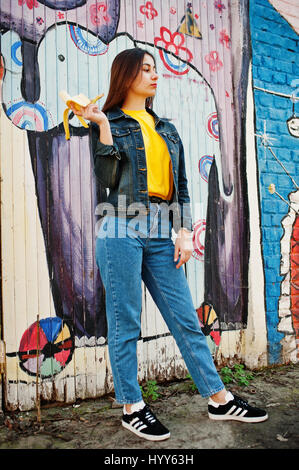 Schöne Teenager-Mädchen essen Bananen, gelbes T-shirt, Jeans in der Nähe von Graffitiwand tragen. Stockfoto