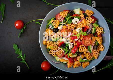 Nudeln in der Form-Herz-Salat mit Tomaten, Gurken, Oliven, Mozzarella und rote Zwiebel griechischen Stil. Flach zu legen. Ansicht von oben Stockfoto