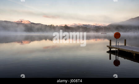 Nebel steigt aus den ruhigen Gewässern des Windermere Lake in Ambleside Pier neben Herbst Wald und unter Thew schneebedeckten Bergen des Langdale, in Engl Stockfoto