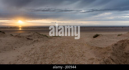 Liverpool, England, UK - 12. November 2016: Die Sonne geht hinter Antony Gormley "Woanders" Skulpturen auf Crosby Strand mit Windparks hinter. Stockfoto