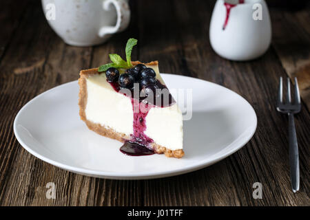 Stück Käsekuchen mit schwarzen Johannisbeeren und Heidelbeeren Sauce auf weißen Teller auf Holztisch Stockfoto