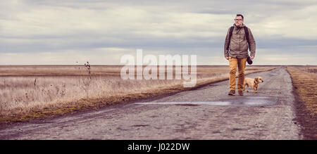 Wanderer und Hund zu Fuß hinunter eine Straße an einem bewölkten Tag Stockfoto