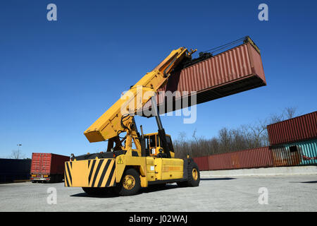 Rückansicht des gelben Containerumschlag Fahrzeug, große rote Container oben in der Luft, niedrigen Winkel Ansicht gegen blauen Himmel Stockfoto