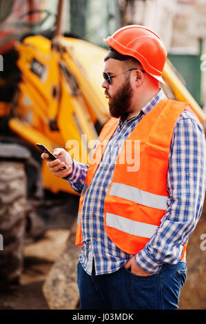 Brutale Bart Arbeiter Mann Anzug Bauarbeiter in orange Helm, Sonnenbrille gegen Traktor mit Mobiltelefon zur hand. Stockfoto