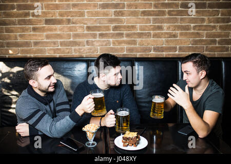 Zeit mit Freunden verbringen. Hübscher junger Mann mit Bier Toasten und lächelnd sitzend mit seinen Freunden im Bier-pub Stockfoto