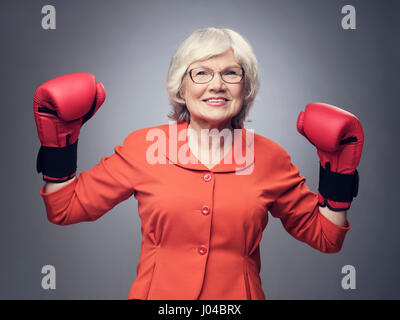 Ältere Dame mit erhobenen Händen in Boxhandschuhen auf grauem Hintergrund lächelt Stockfoto