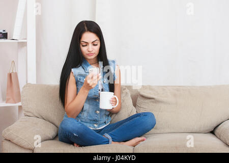 Junge Frau, Lesen von Nachrichten mobile während Tasse Tee und sitzen auf couch Stockfoto