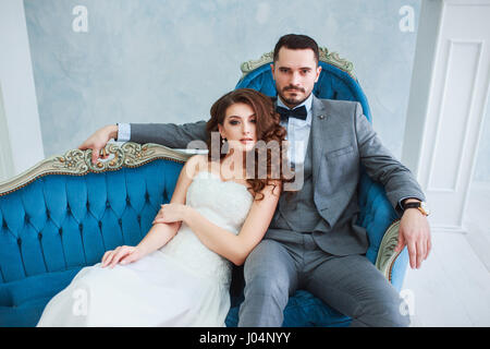 Braut im Kleid und der Bräutigam im grauen Anzug auf Sofa im Innenbereich in weisse Studio Interieur wie zu Hause zu sitzen. Stockfoto