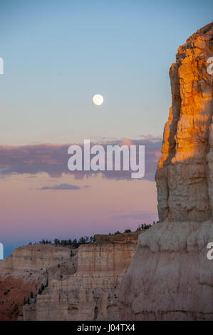 Vollmond wird neben einer senkrechten Felswand Rost durch eine untergehende Sonne gefärbt.  Schichten der rosa Wolke ergänzen dieses vertikale Bild. Stockfoto
