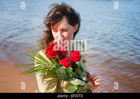 Traumhafte Frau mit roten Rosen gegen das Meer im sonnigen Tag. Brünette in einem Demi-Saison, nach draußen. Dating, Romantik und Erinnerungen Thema Stockfoto