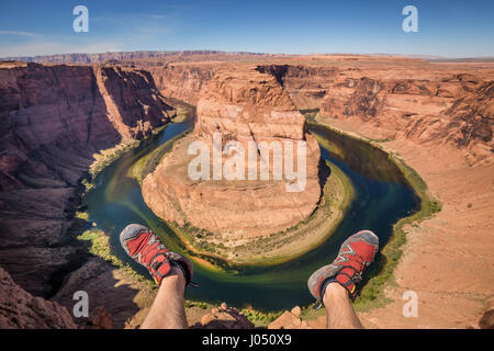 Im Weitwinkel ein junger Wanderer hängen seine Füße von einer Klippe am berühmten Horseshoe Bend an einem schönen sonnigen Tag mit blauem Himmel und Wolken, Arizona, USA Stockfoto