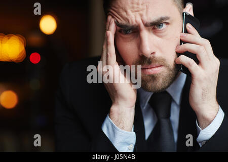Porträt des modernen bärtigen Geschäftsmann sucht verwirrt und frustriert während des Gesprächs mit dem Handy mit Problem-Client gegen schwarze Blendenfleck zurück Stockfoto