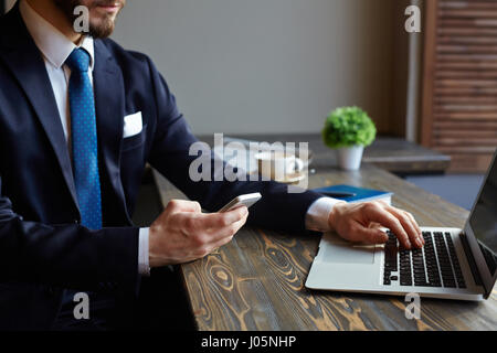 Nahaufnahme des eleganten modernen Geschäftsmann in Kaffeepause: mit Smartphone und Eingabe von SMS-Nachrichten während der Arbeit mit Laptop im Café Stockfoto