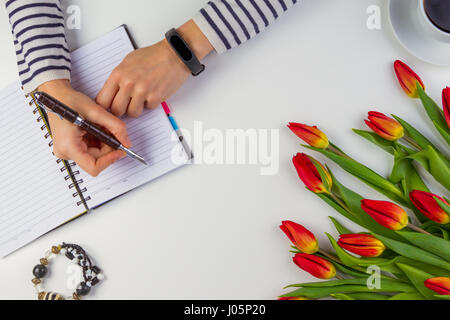 Frau Hand schreibt auf Papier-Notizbuch mit Stift. Wunderschöne Blumen auf weißen Tisch. Stockfoto